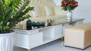 A modern living room with a white sofa, a glass coffee table, and a beige ottoman. There is a potted plant on the left and a vase with red roses on the right. - Sea View Hotel Phan Thiet (Tien Thanh)