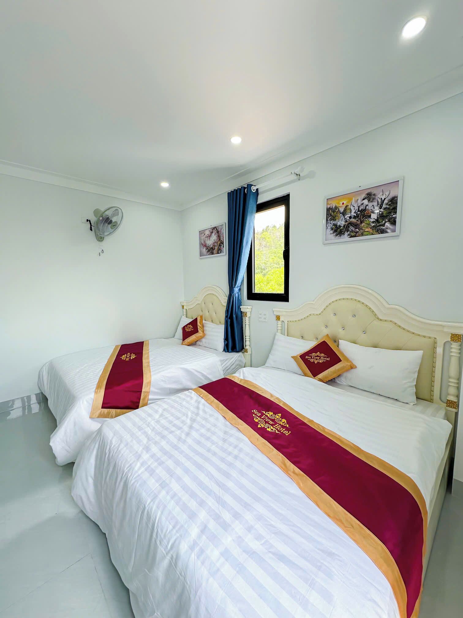 A hotel room with two beds, each with white bedding and maroon accents. There is a ceiling fan, a window with blue curtains, and two wall-mounted paintings.