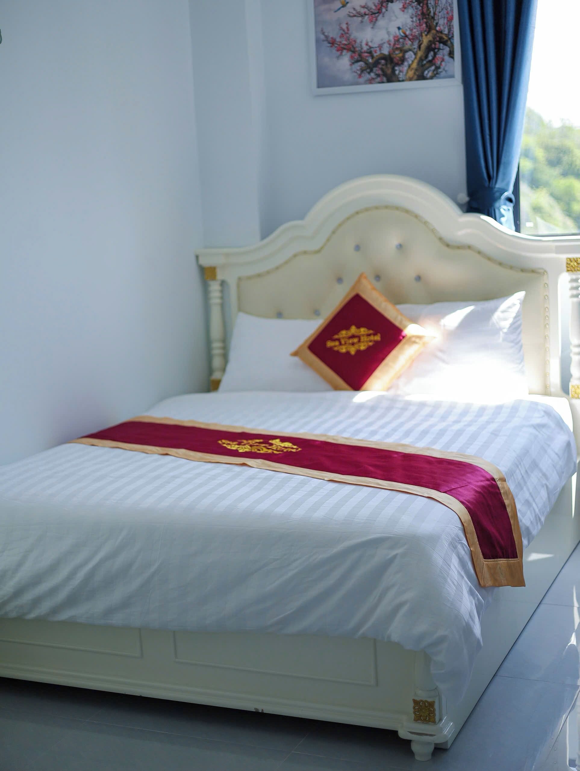 Una cama bien hecha con cabecera blanca, un cojín decorativo rojo y dorado, y una colcha blanca con ribete dorado.