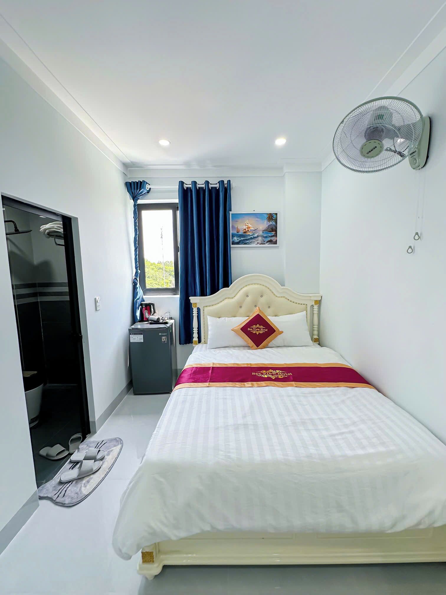 Une chambre d’hôtel avec un lit, un ventilateur de chevet, une fenêtre avec des rideaux, un petit bureau et une vue sur l’extérieur.
