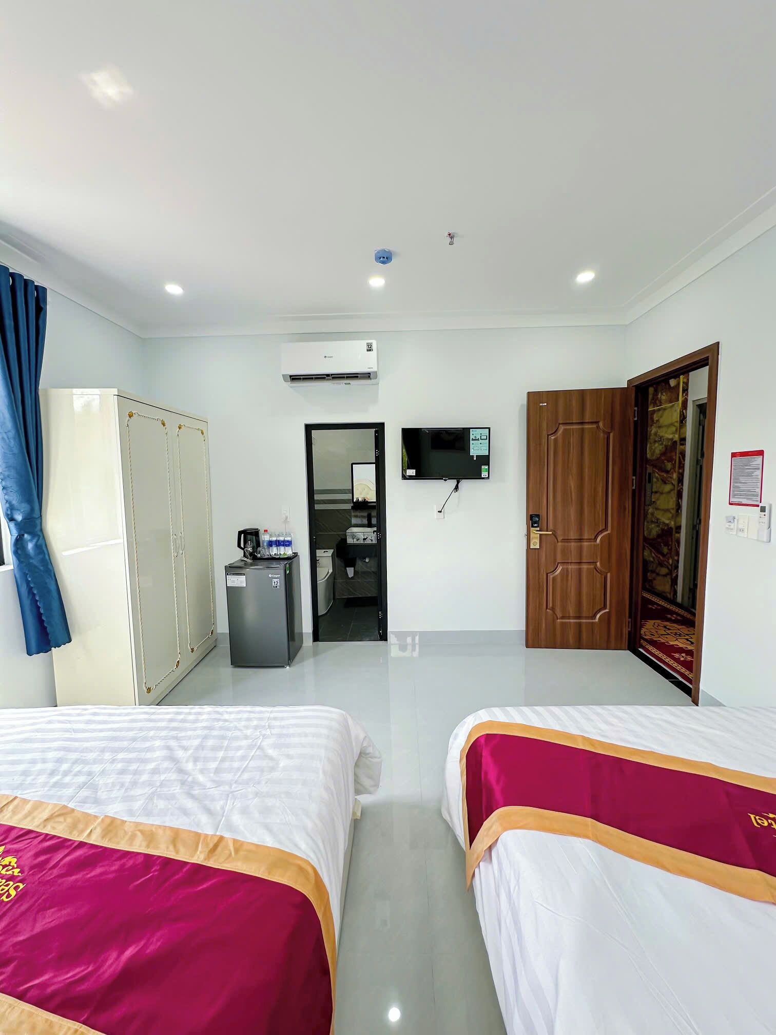 A hotel room with two beds, a wardrobe, a desk with a computer, an air conditioning unit, a television, and a bathroom door.