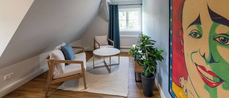 A modern loft bedroom with a sloped ceiling, a cozy seating area with a sofa and a small table, a potted plant, and a vibrant mural on the wall.
