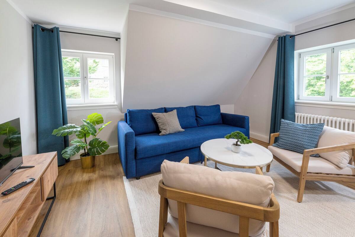 A modern living room with a blue sofa, a round coffee table, a beige armchair, and a wooden TV stand. There are two potted plants, one near the sofa and one near the coffee table.