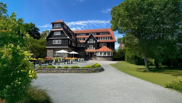A large, multi-story building with a stone facade, a red-tiled roof, and a paved driveway leading to a seating area with umbrellas. - Wohnpark Hotel (Heppenheim)