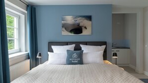 A hotel room with a bed, a nightstand, a painting of a beach scene, and a kitchenette. - Wohnpark Hotel (Heppenheim)