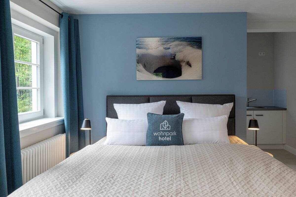 A hotel room with a bed, a nightstand, a painting of a beach scene, and a kitchenette.