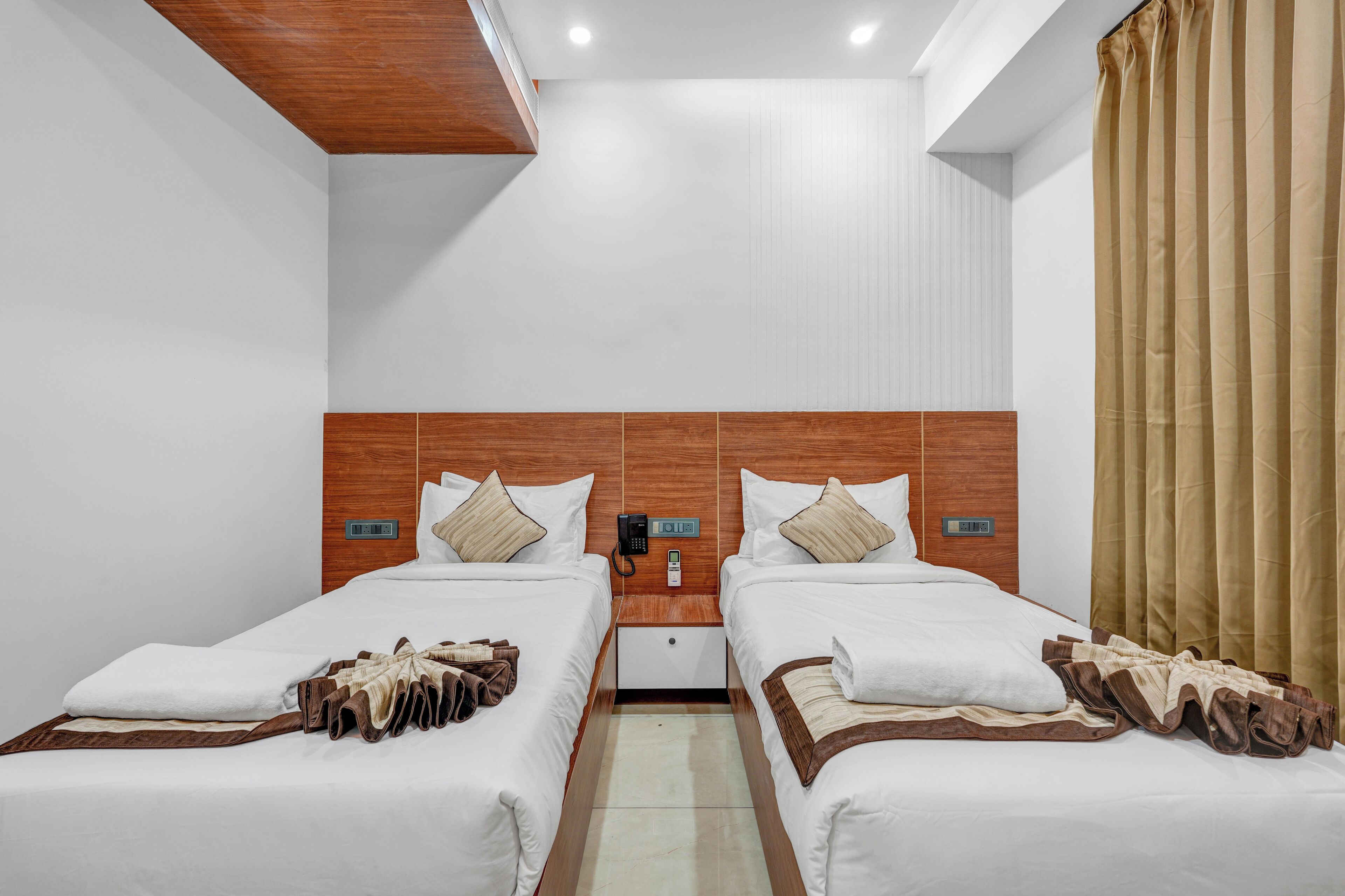 Ein Hotelzimmer mit zwei Betten, einem Holz-Kopfende, weißen Wänden, beigen Vorhängen und einem kleinen Nachttisch mit Telefon.