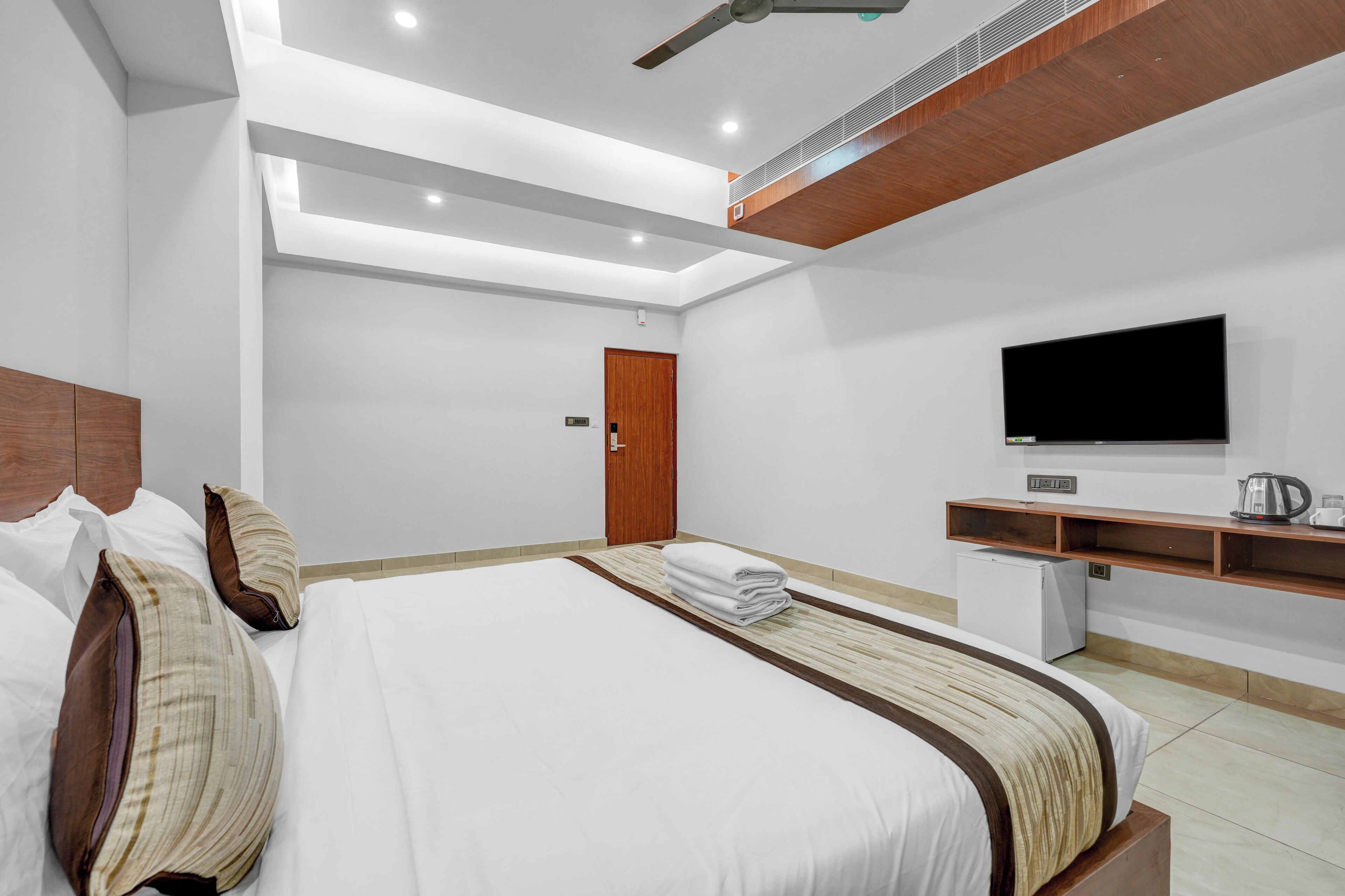 A modern hotel room with a large bed, a flat-screen TV, a small kitchenette, and a wooden ceiling.