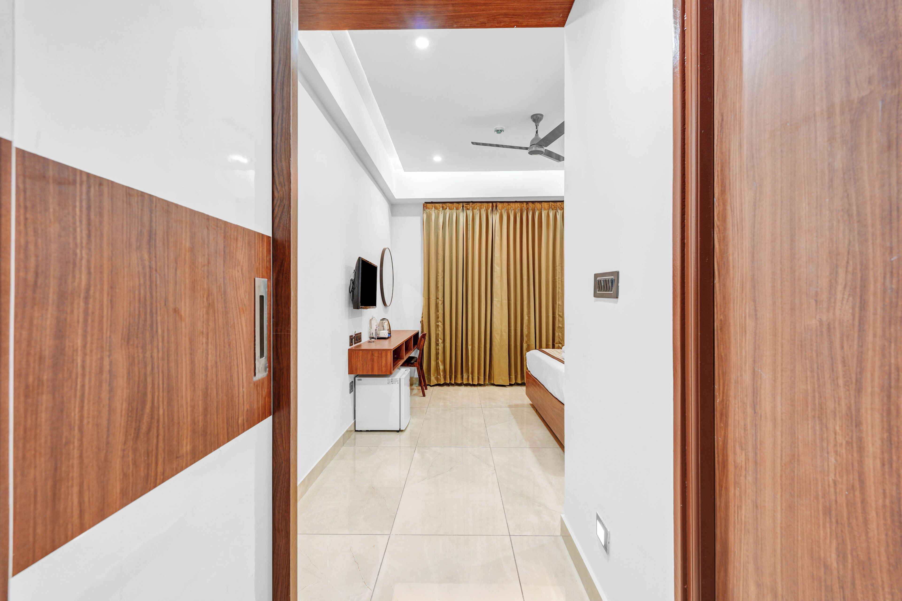 Une chambre d’hôtel avec une armoire en bois, un bureau avec une chaise, un petit réfrigérateur, un lit et un ventilateur de plafond.