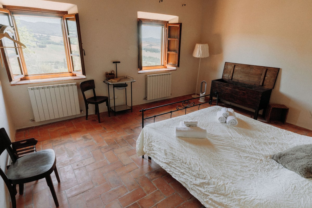 Een slaapkamer met een bed, een stoel, een bureau, een lamp en twee ramen.