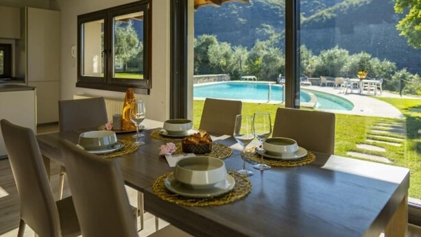A dining table set with plates, glasses, and a cake, overlooking a swimming pool and lush greenery. - Villa Anna Lake View (Pilzone)