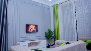 A modern living room with a flat-screen TV, a white entertainment unit, a beige sofa, and a green plant. - Cozy Residence I (Kampala)