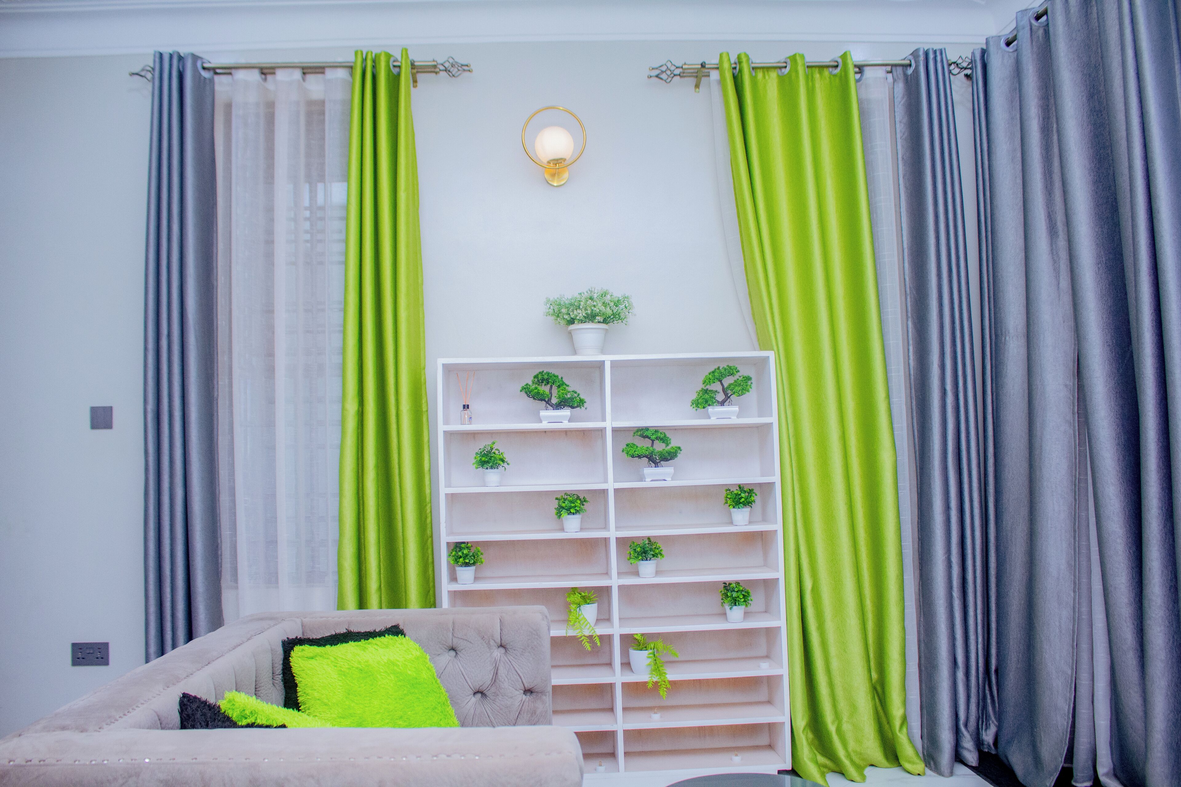 Una sala de estar con un sofá, una estantería blanca con plantas y cortinas verdes.