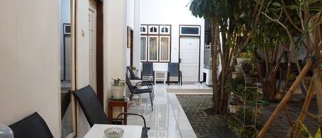 A narrow corridor with a tiled floor leading to a door marked '9'. There are black chairs and a small table with a glass bowl on it. The corridor is flanked by walls with windows and a potted plant on the right.