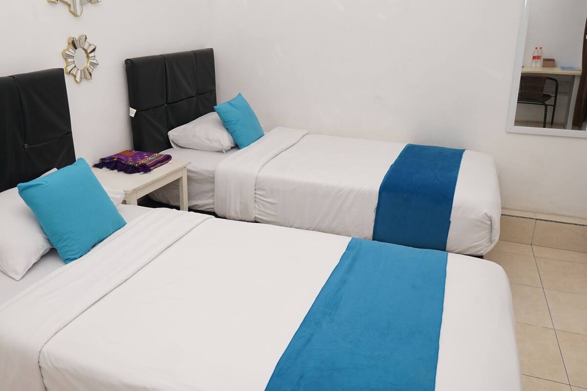 Habitació d'hotel amb dos llits, cadascun amb un coixí blau decoratiu i un llençol blanc. Hi ha una taula petita amb una manta morada i un mirall a la paret.