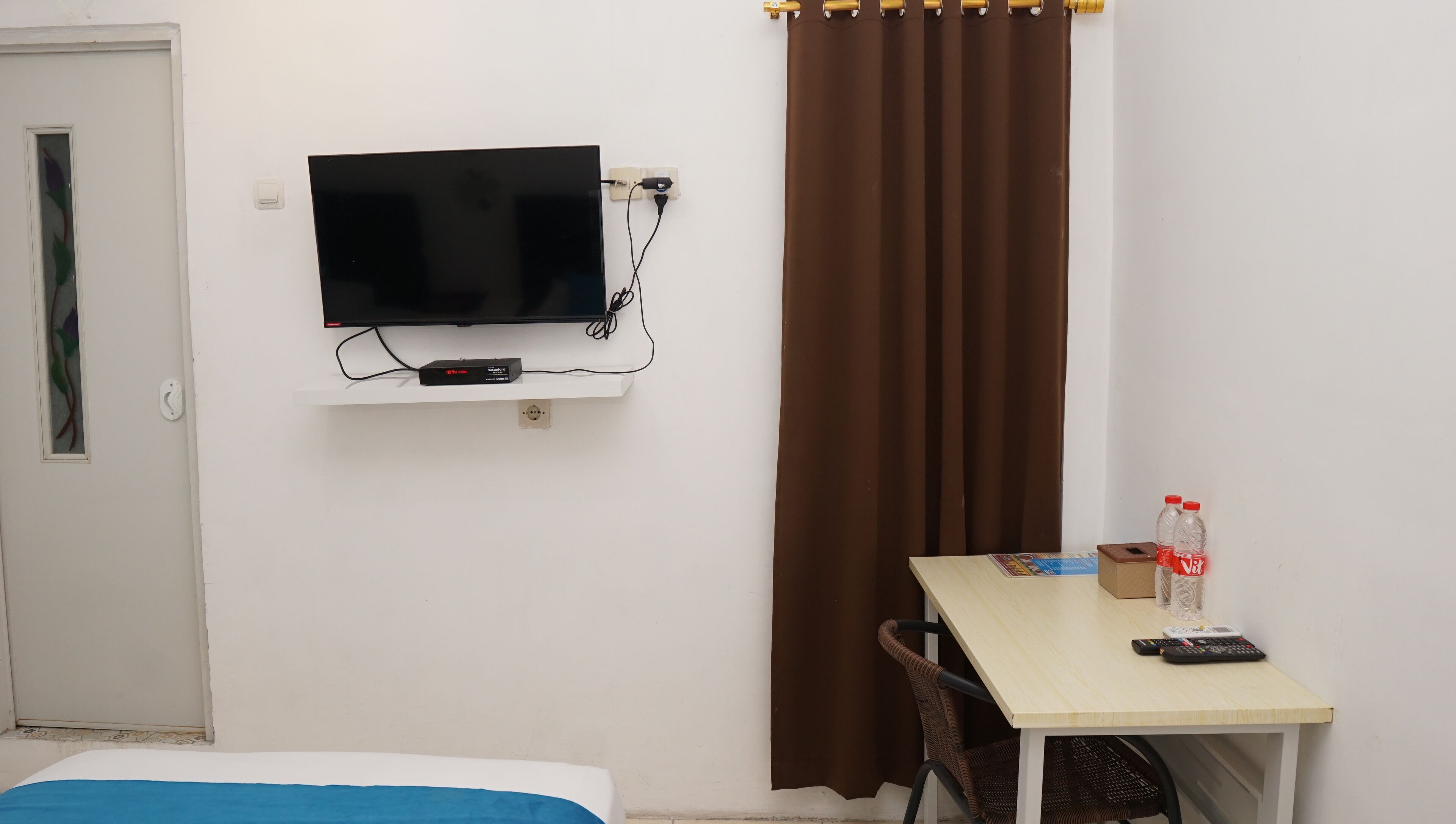 A hotel room with a bed, a TV mounted on the wall, a table with a remote control, a bottle of water, and a brown curtain.