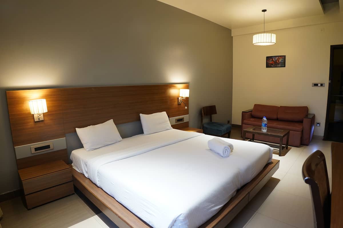 Una habitación de hotel con una cama grande, lámparas de noche, cabecero de madera, una mesita pequeña, un sofá e imagen colgada en la pared.
