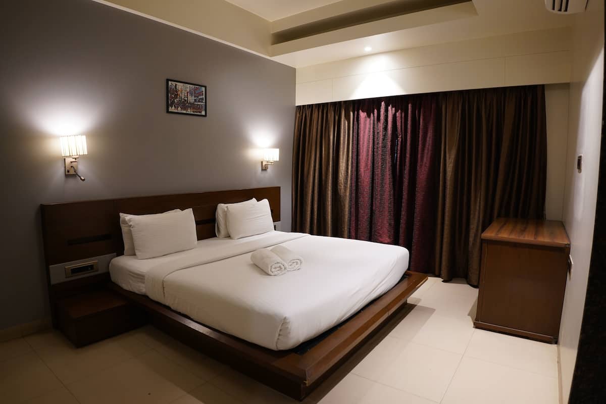 Una habitación de hotel con una cama grande, dos lámparas de noche, una mesita de noche, un cabecero de madera, un armario pequeño y una ventana con cortinas.
