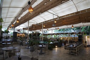 An indoor restaurant with high ceilings, hanging lights, and a variety of seating arrangements including tables and chairs. - Hotel Sadanand Regency - Balewadi, Baner (Paud)