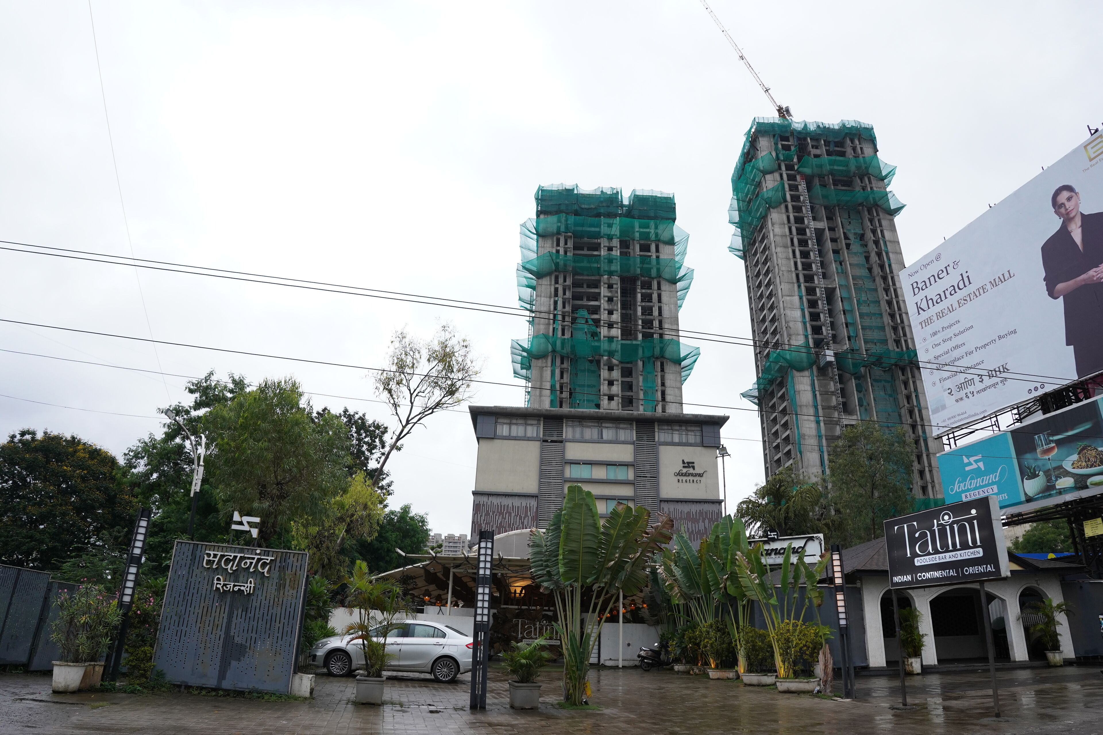 A multi-story building with a sign for 'Sadanand Regency' and a partially constructed building with green netting.
