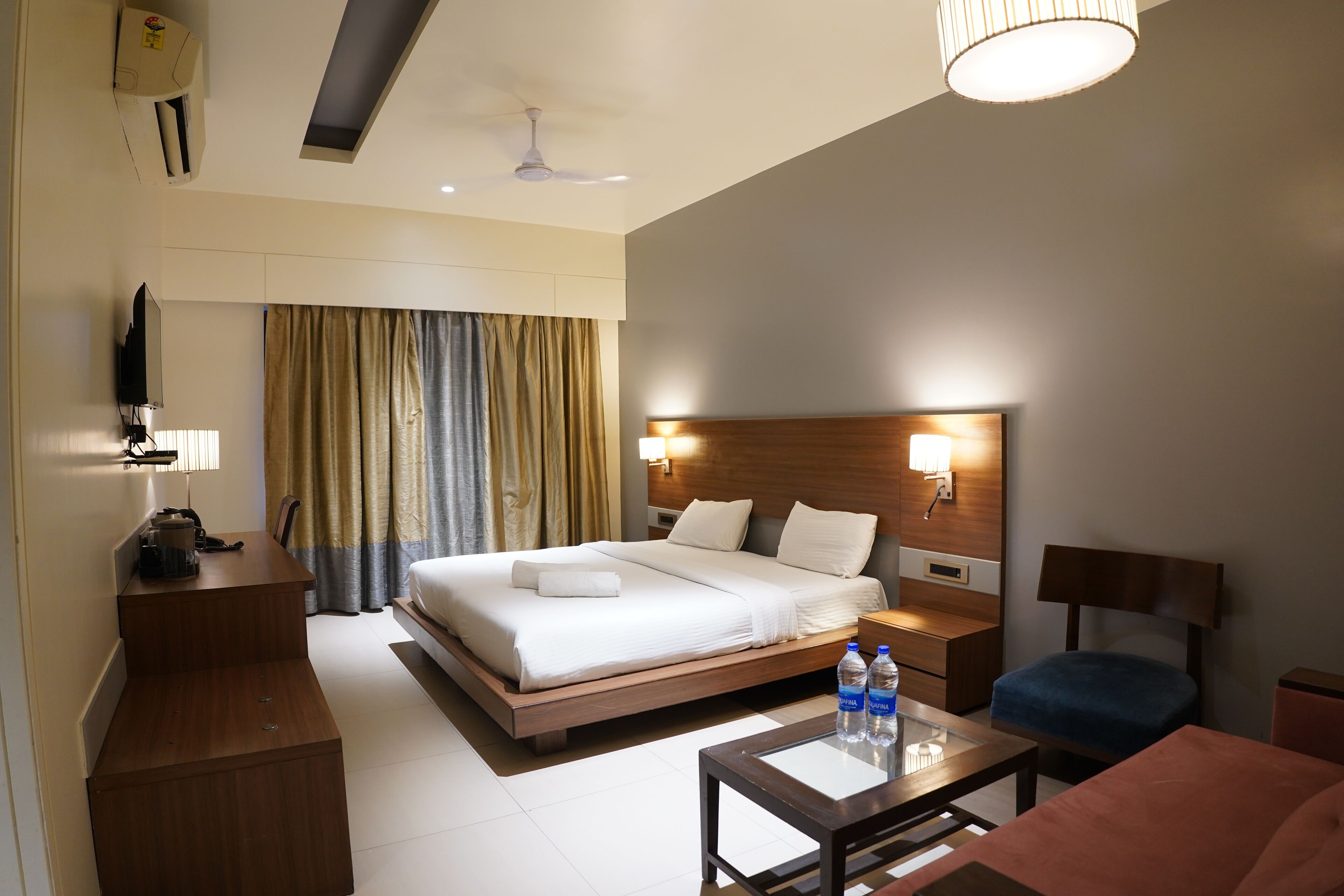 A hotel room with a large bed, bedside tables, a desk, a chair, a TV, and a ceiling fan.