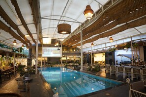 An indoor swimming pool with a ceiling that has a unique woven design, surrounded by tables and chairs. - Hotel Sadanand Regency - Balewadi, Baner (Paud)