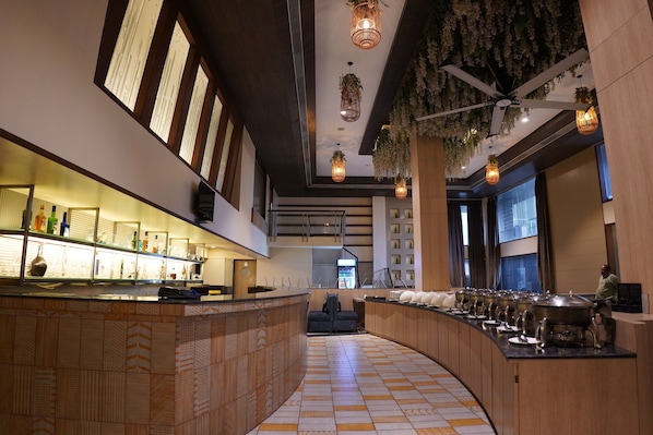 Zona de bar moderna con barra curva, luces colgantes y techo con ventilador y plantas.