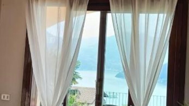 Quarto com uma grande janela coberta por cortinas brancas translúcidas, com vista para um corpo d'água e montanhas ao longe.