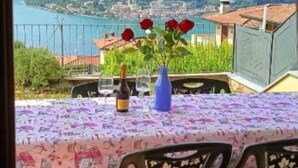 Uma mesa com toalha florida, uma garrafa e um vaso com flores vermelhas, com vista para uma cidade costeira com edifícios e vegetação.