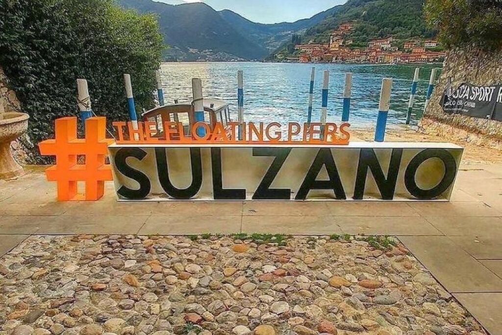 A sign with the text "The Floating Pier #SULZANO" and a view of a coastal area with buildings and water.
