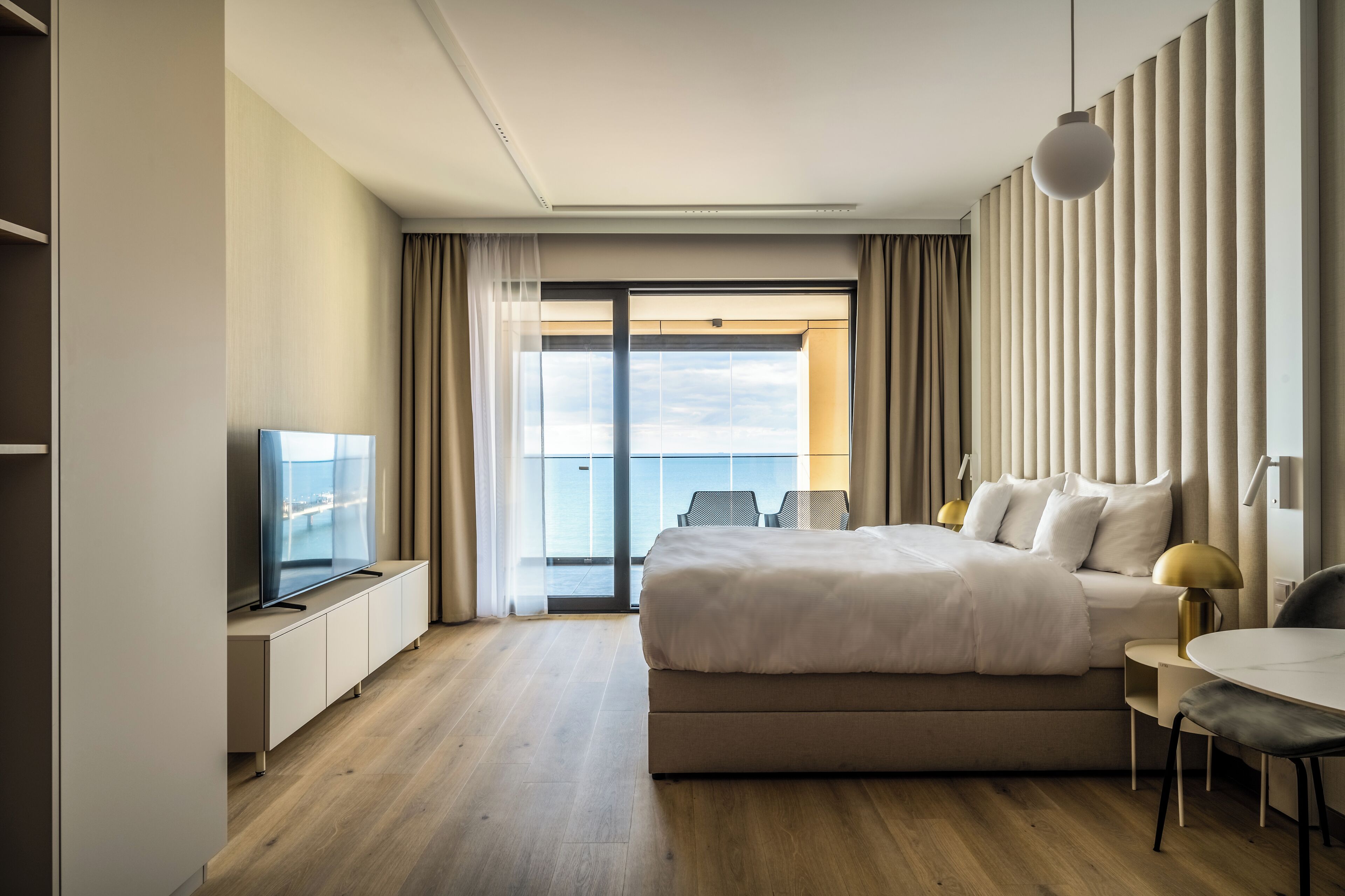 A modern hotel room with a large bed, a TV, a balcony with curtains, and a view of the sky.