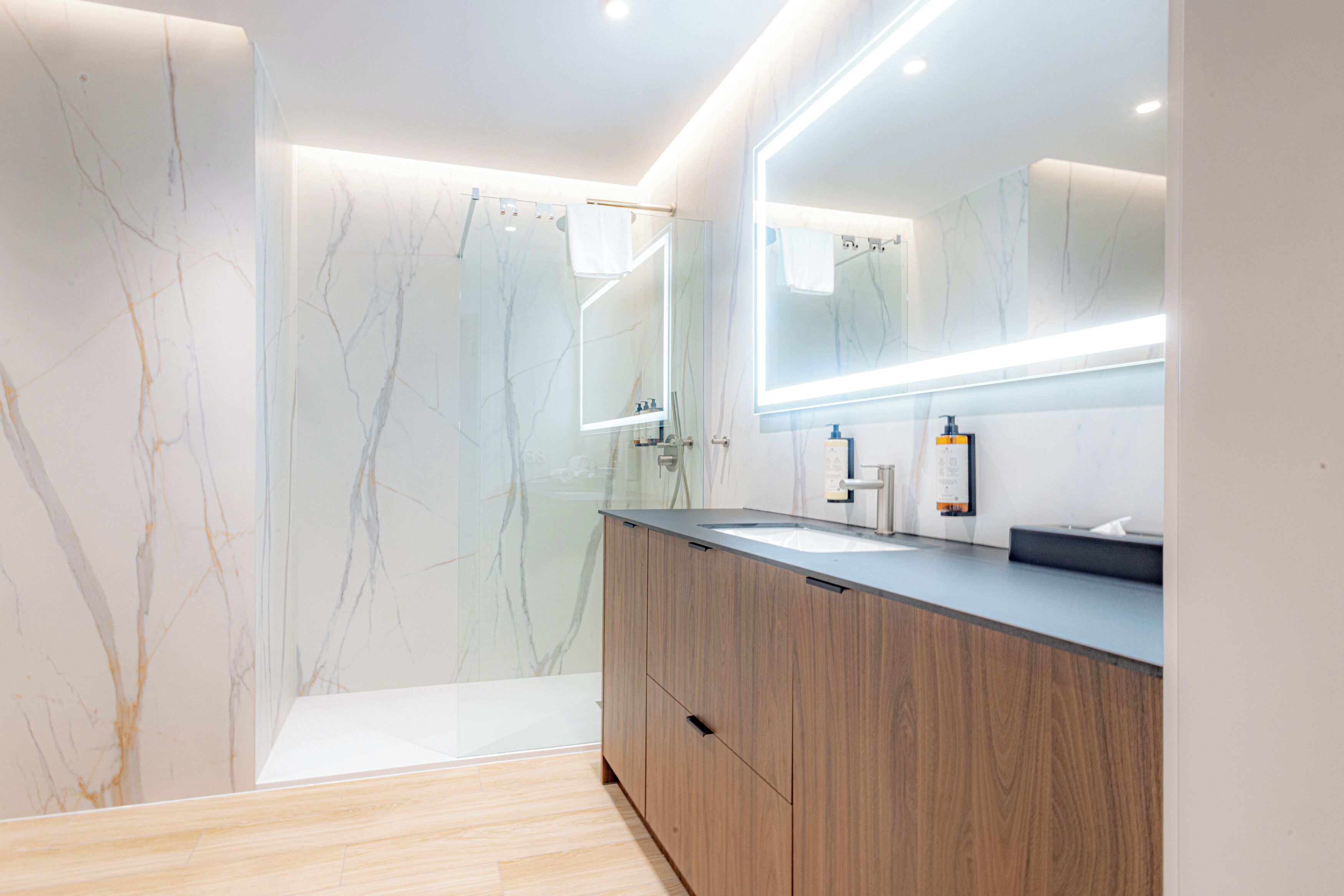 A modern bathroom with a marble wall, wooden vanity, and a large mirror.
