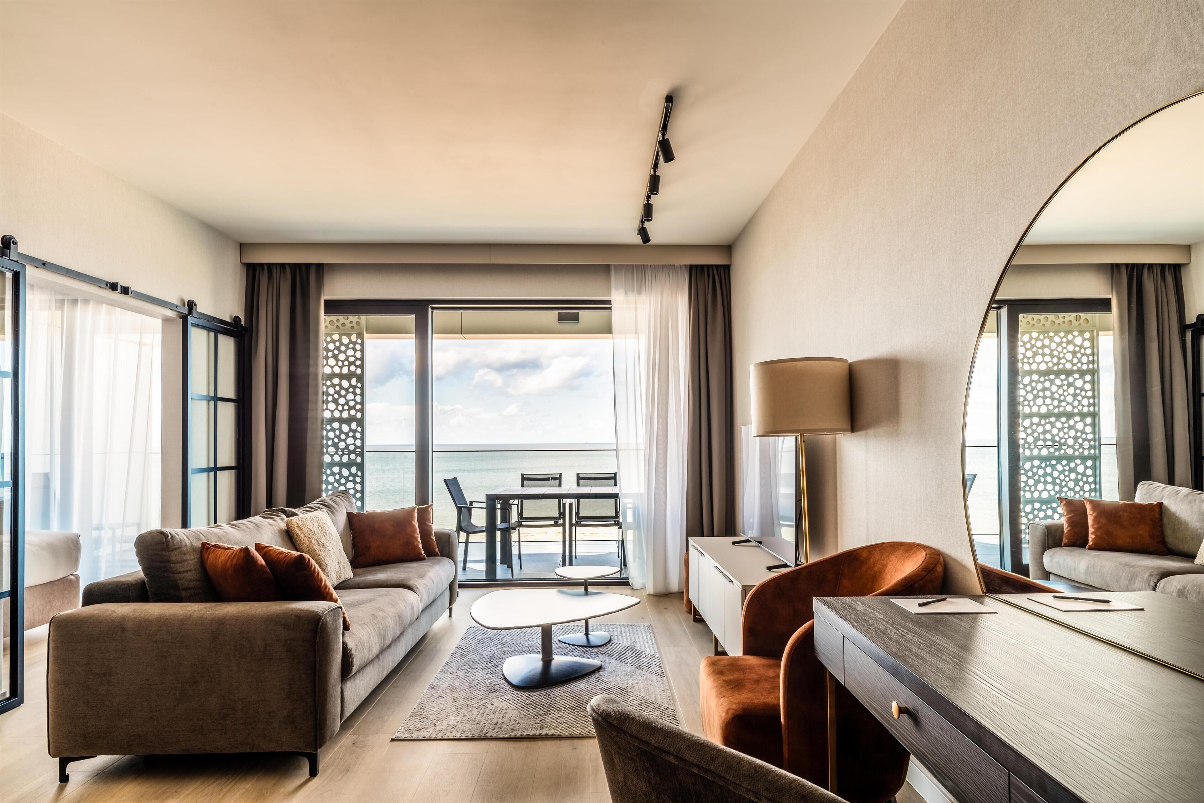 Ein modernes Hotelzimmer mit einer Couch, einem Sessel, einem Esstisch und Meerblick durch große Fenster.