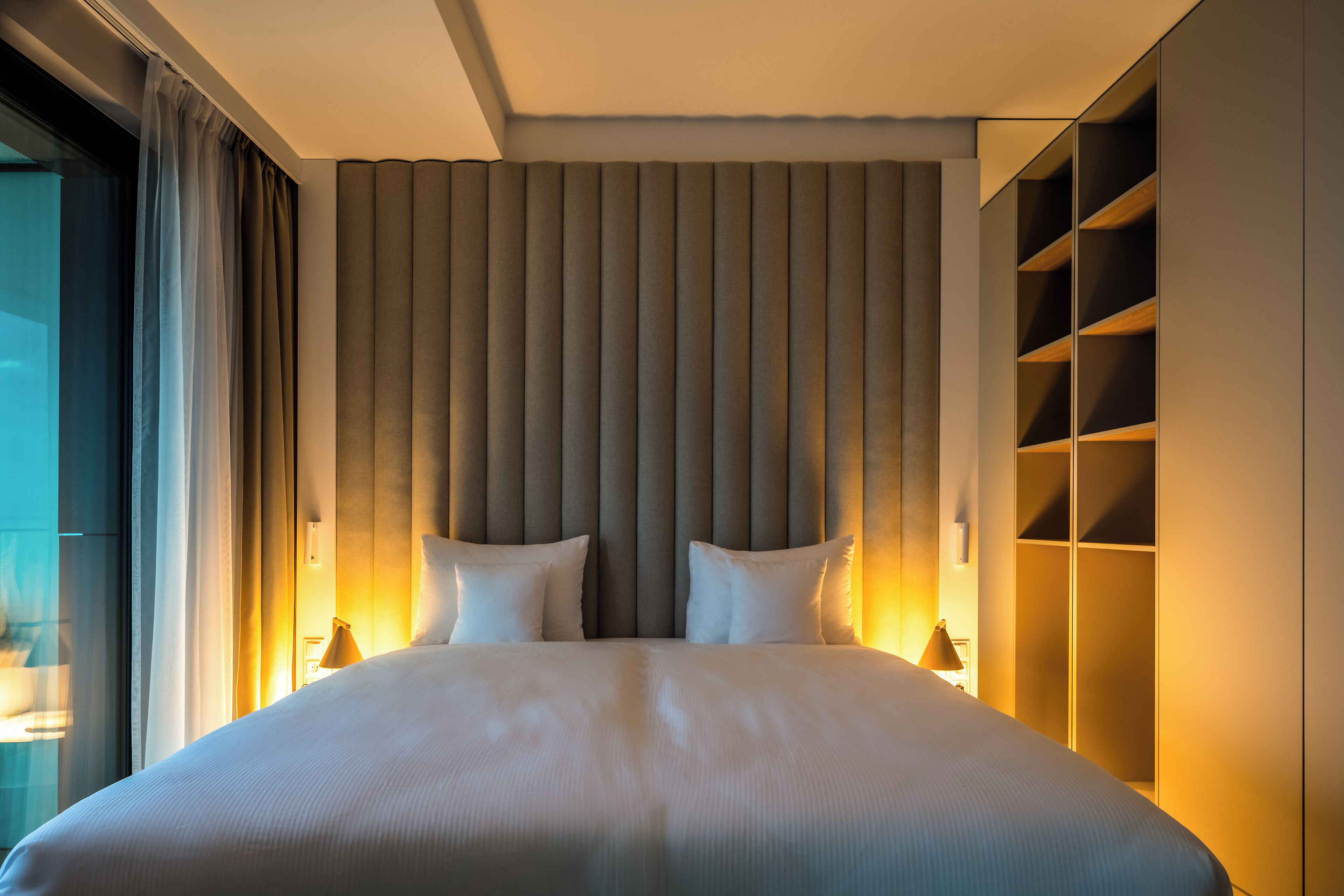 Una habitación de hotel moderna con una cama grande, un cabecero con líneas verticales y estanterías empotradas a la derecha.