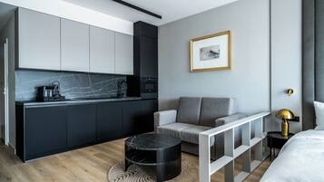 Une chambre d’hôtel moderne dotée d’un coin cuisine, d’un canapé gris, d’une table basse ronde noire et d’un lit avec une literie blanche.
