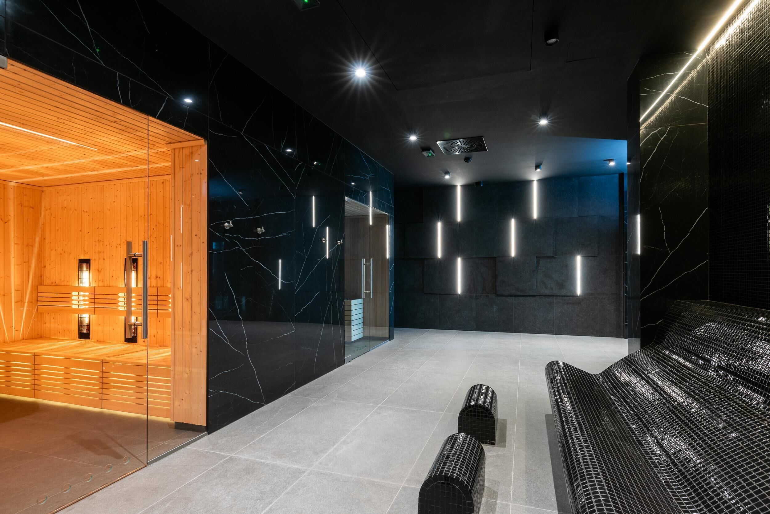 Un espace spa moderne avec des sièges en bois, une cloison en verre et un mur en marbre foncé agrémenté de luminaires.