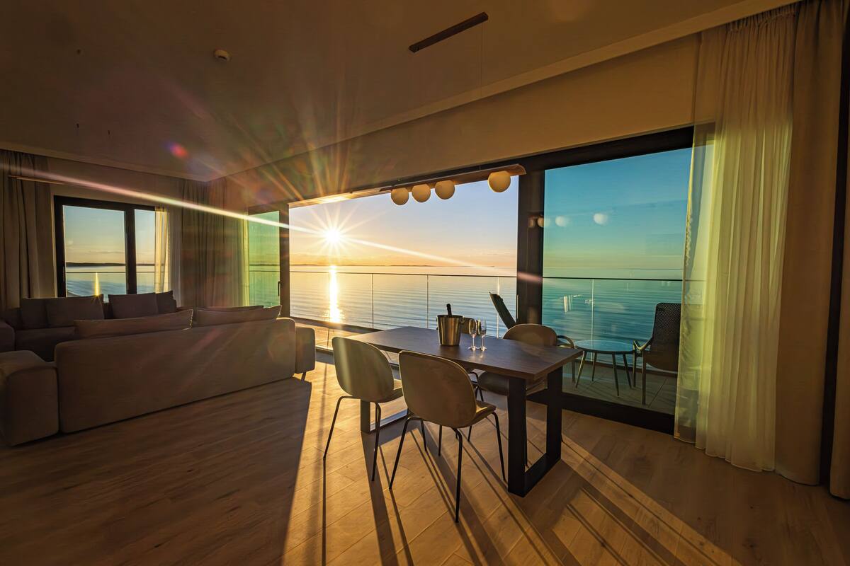 Un salon moderne avec un plancher en bois, un canapé, une table à manger avec des chaises et une grande fenêtre offrant une vue sur l’océan au coucher du soleil.