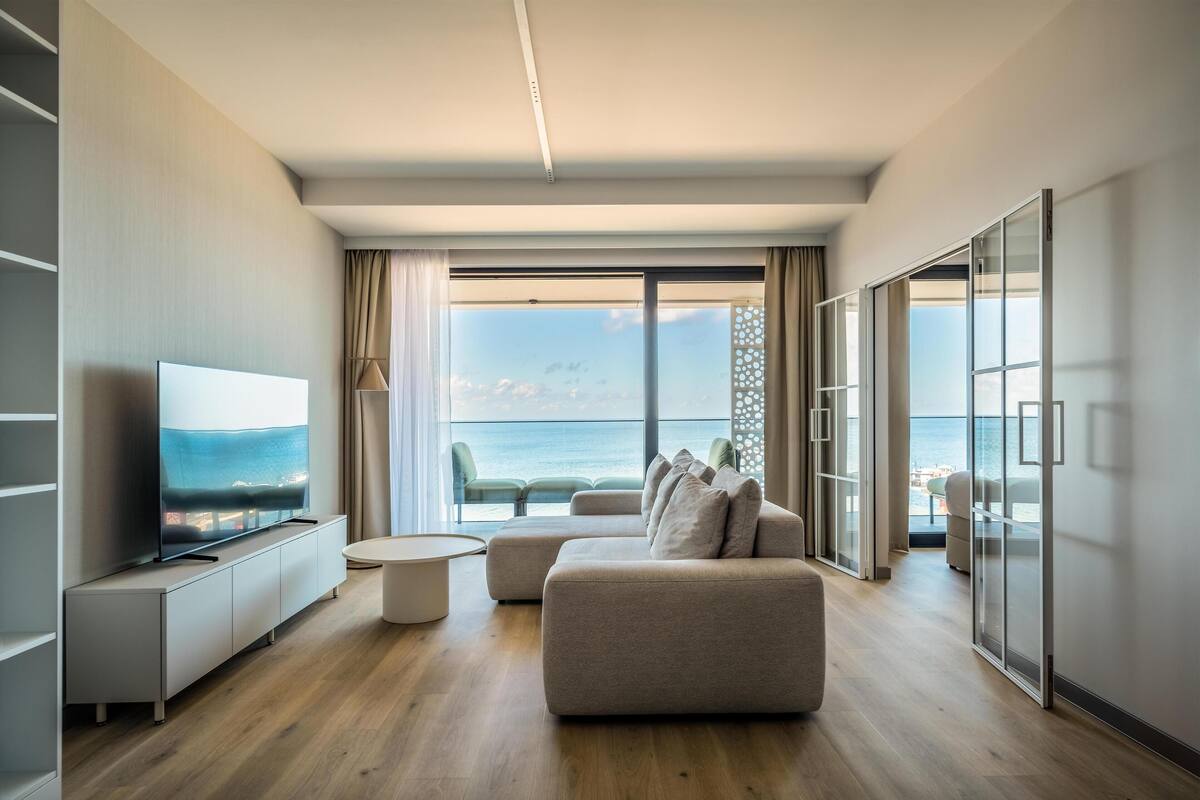 Un salon moderne doté d’un grand téléviseur à écran plat, d’un canapé et d’une table basse ronde. Un balcon offre une vue sur la mer.