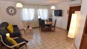 A living room with a sofa, a dining table with chairs, a clock, and a TV on the wall. - Pavilion well located for visiting the region (Ruelisheim)