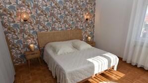 A bedroom with a floral wallpaper, a wooden floor, a bed with a white bedspread, two bedside lamps, and a wicker nightstand. - Pavilion well located for visiting the region (Ruelisheim)