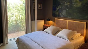 Une chambre à coucher avec un lit, une table de chevet, une lampe, un tableau et une vue sur la verdure par la fenêtre.