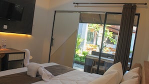 A hotel room with a bed, a desk with a chair, a TV mounted on the wall, and a sliding glass door leading to an outdoor area with plants and furniture. - The Kantun Ungasan (Ungasan)