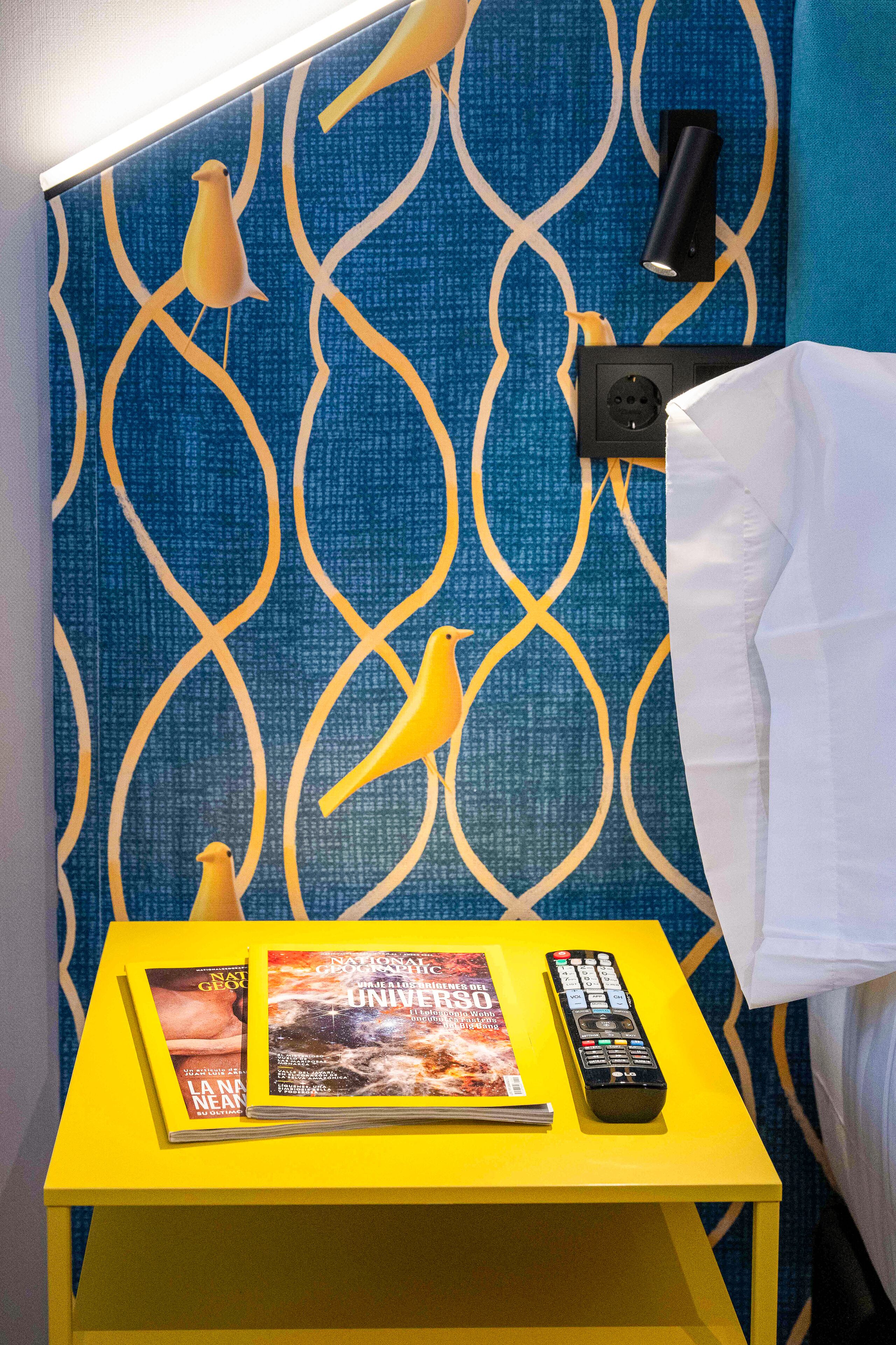 Une table jaune avec des magazines et une télécommande, un mur bleu à motifs et un rideau blanc.