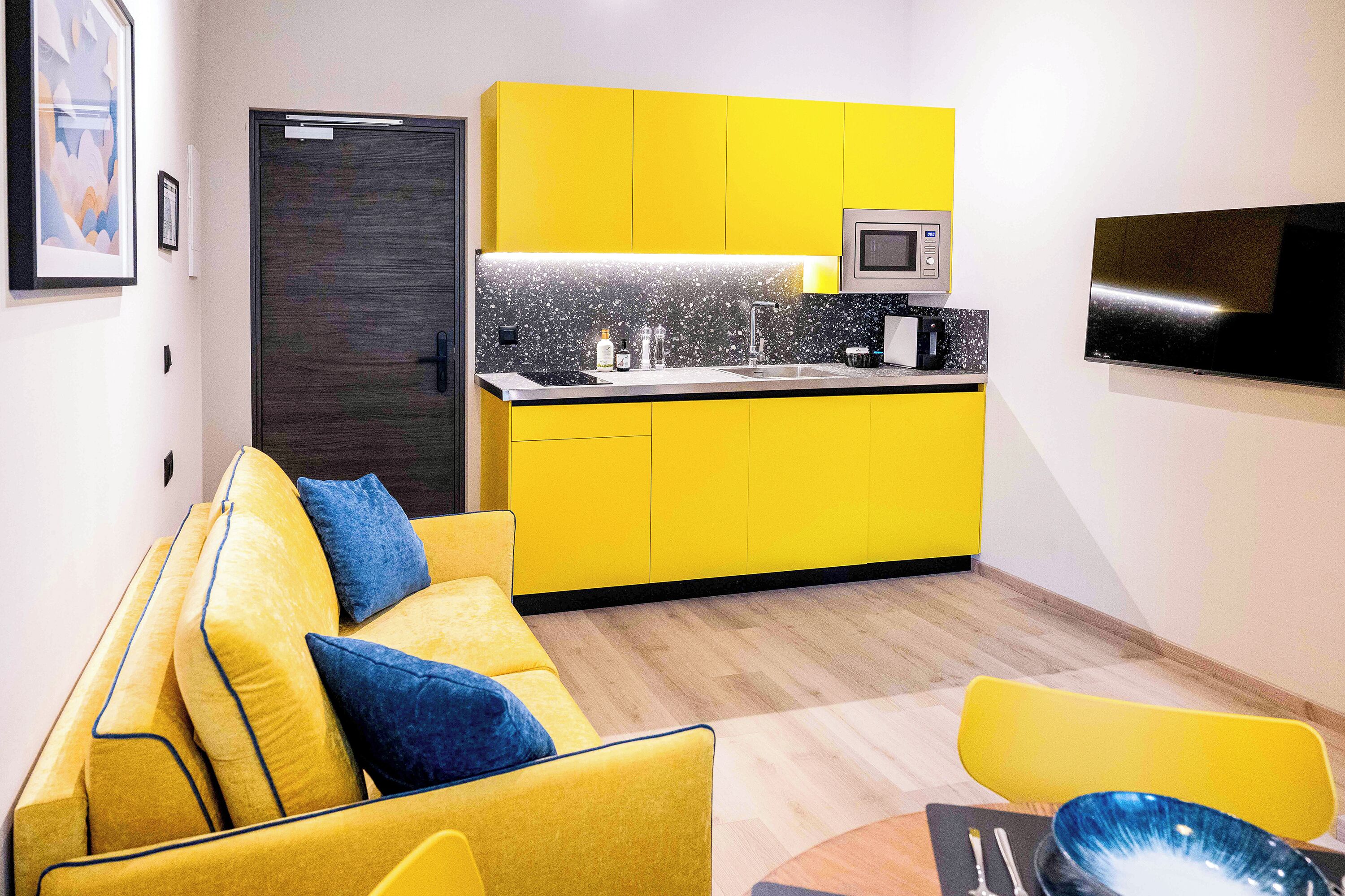 Un espacio habitable compacto con un sofá amarillo, una pequeña área de cocina con microondas y un televisor de pantalla plana montado en la pared.