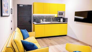 Un espacio habitable compacto con un sofá amarillo, una pequeña área de cocina con microondas y un televisor de pantalla plana montado en la pared.