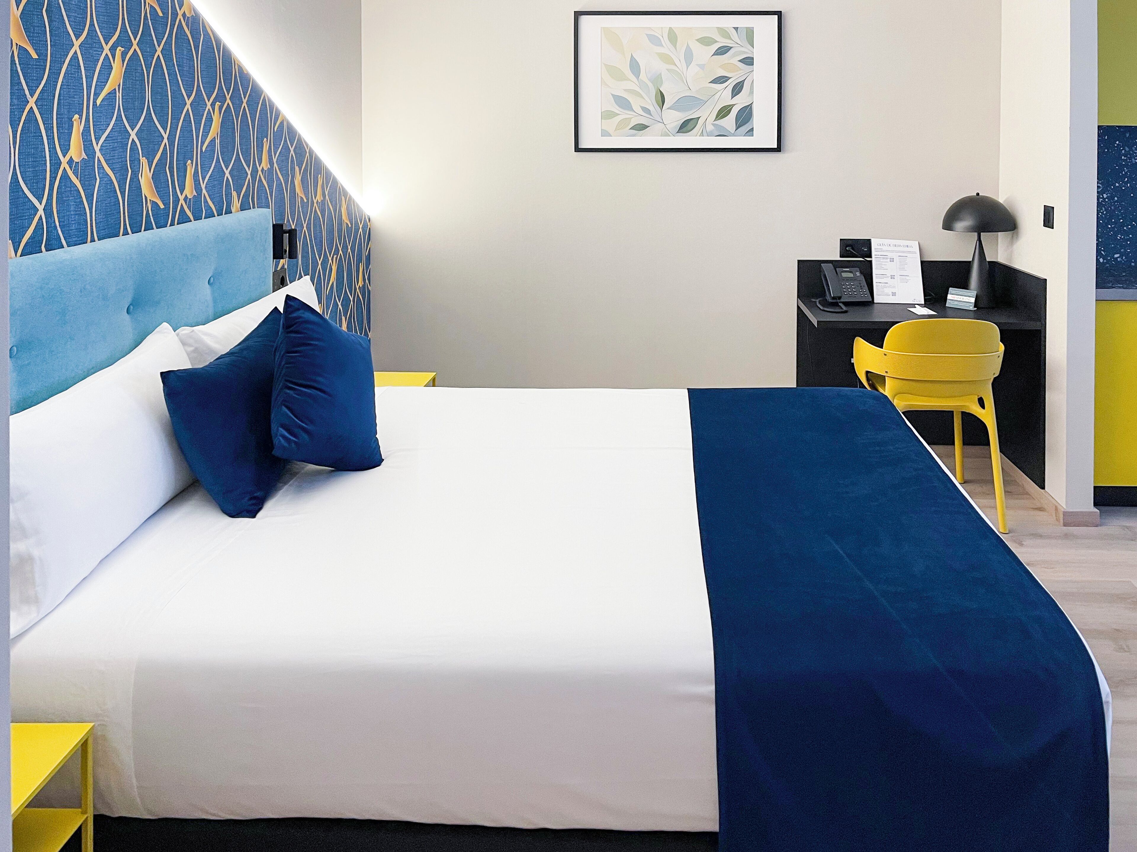 Quarto de hotel com uma cama, uma escrivaninha, uma cadeira e um quadro na parede.