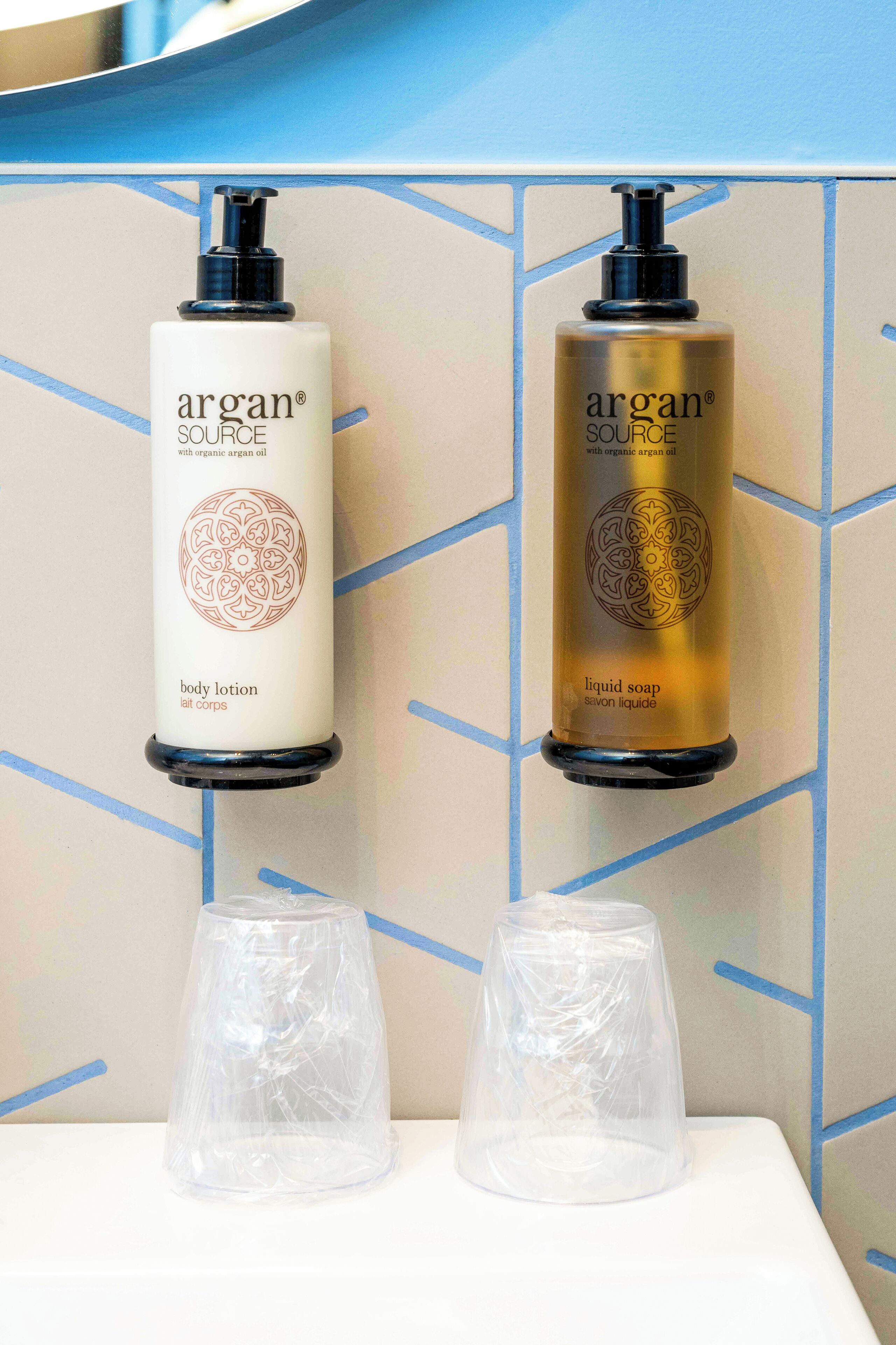 Two Argan Source bottles, one body lotion and one liquid soap, on a wall with a geometric pattern.