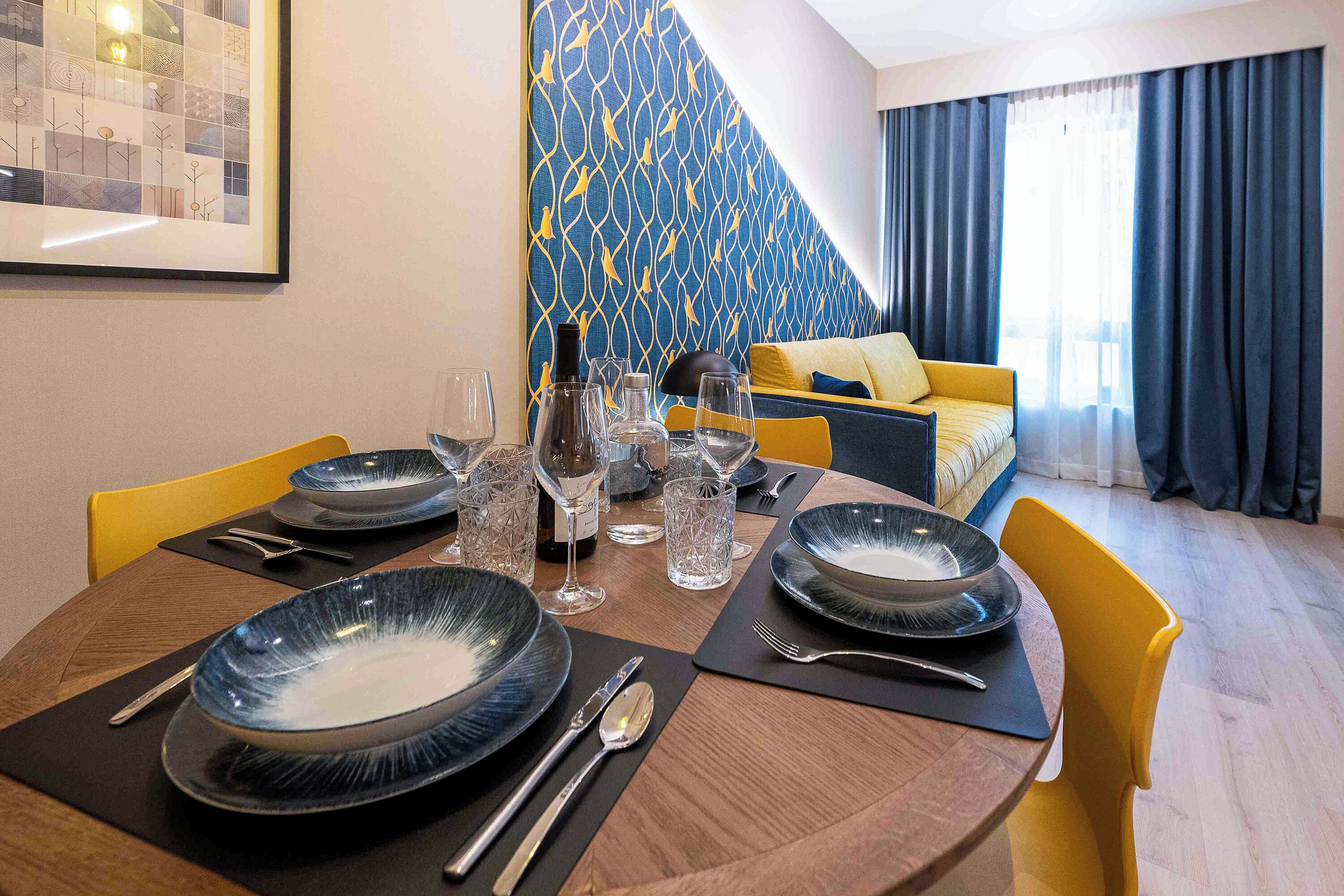 Een eetgedeelte met een ronde tafel voor vier personen, met blauwe borden en bestek. Een gele bank en een behang met patroon zijn op de achtergrond te zien.