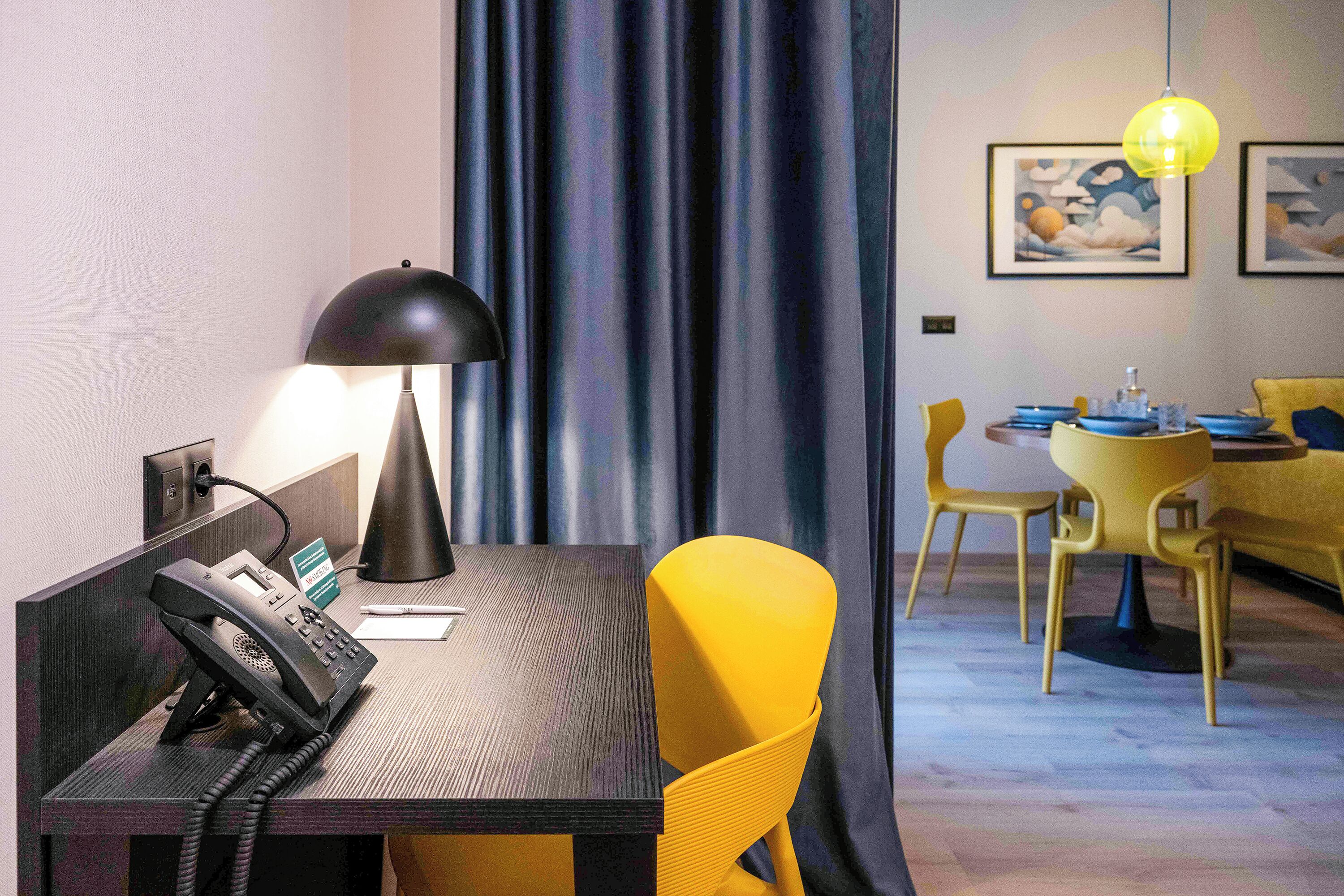 A hotel room with a desk, a telephone, a lamp, a chair, a dining table with chairs, and artwork on the wall.