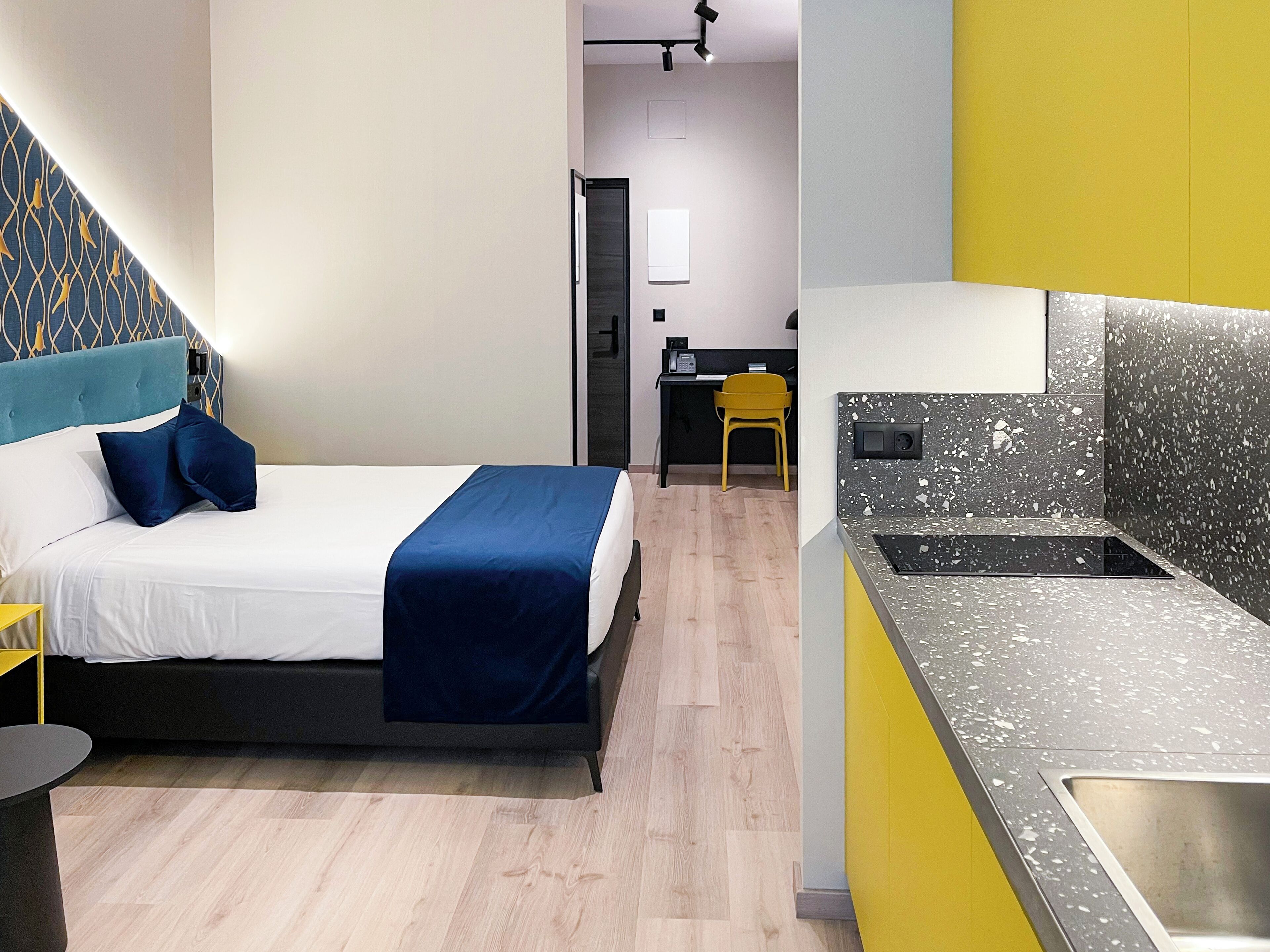 A modern hotel room with a bed, a small desk, and a kitchenette.